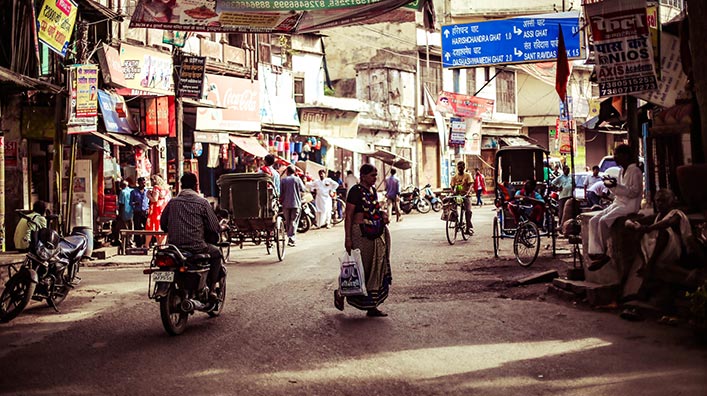 A street with many small shops. A woman crosses the street as cyclists and wagons pass by.